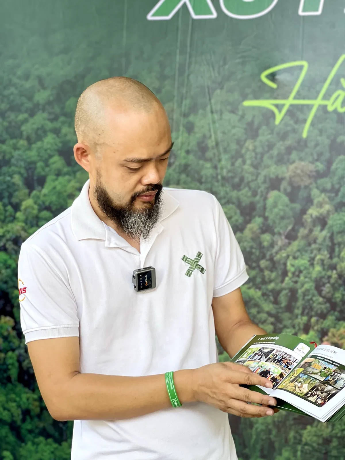 Phạm Thanh Tùng lật xem trang sách có nhiều ảnh màu, đứng trước phông rừng cây xanh