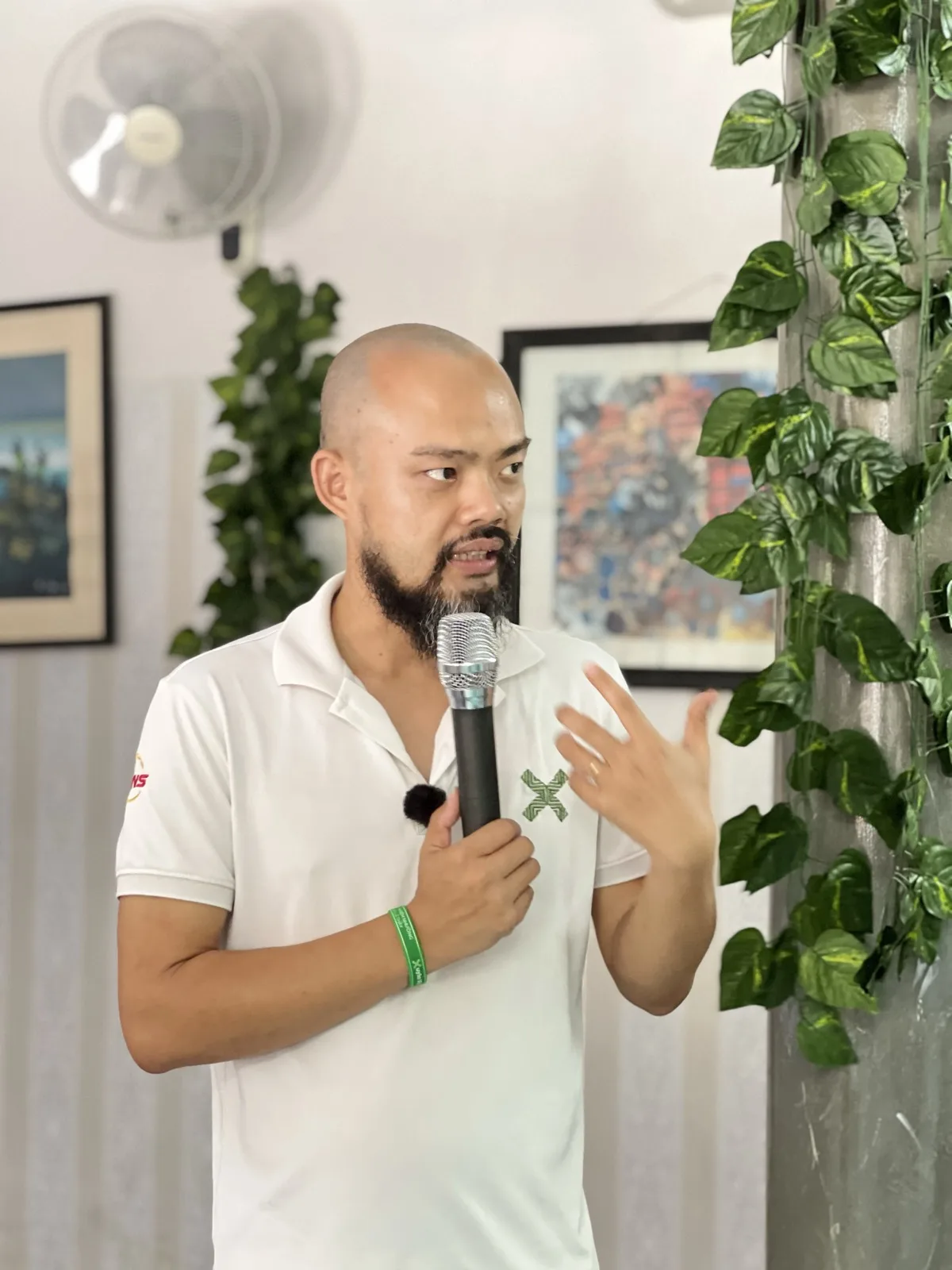 Cận cảnh Phạm Thanh Tùng cầm micro, tay phải xòe ra khi giải thích, cạnh giàn lá xanh