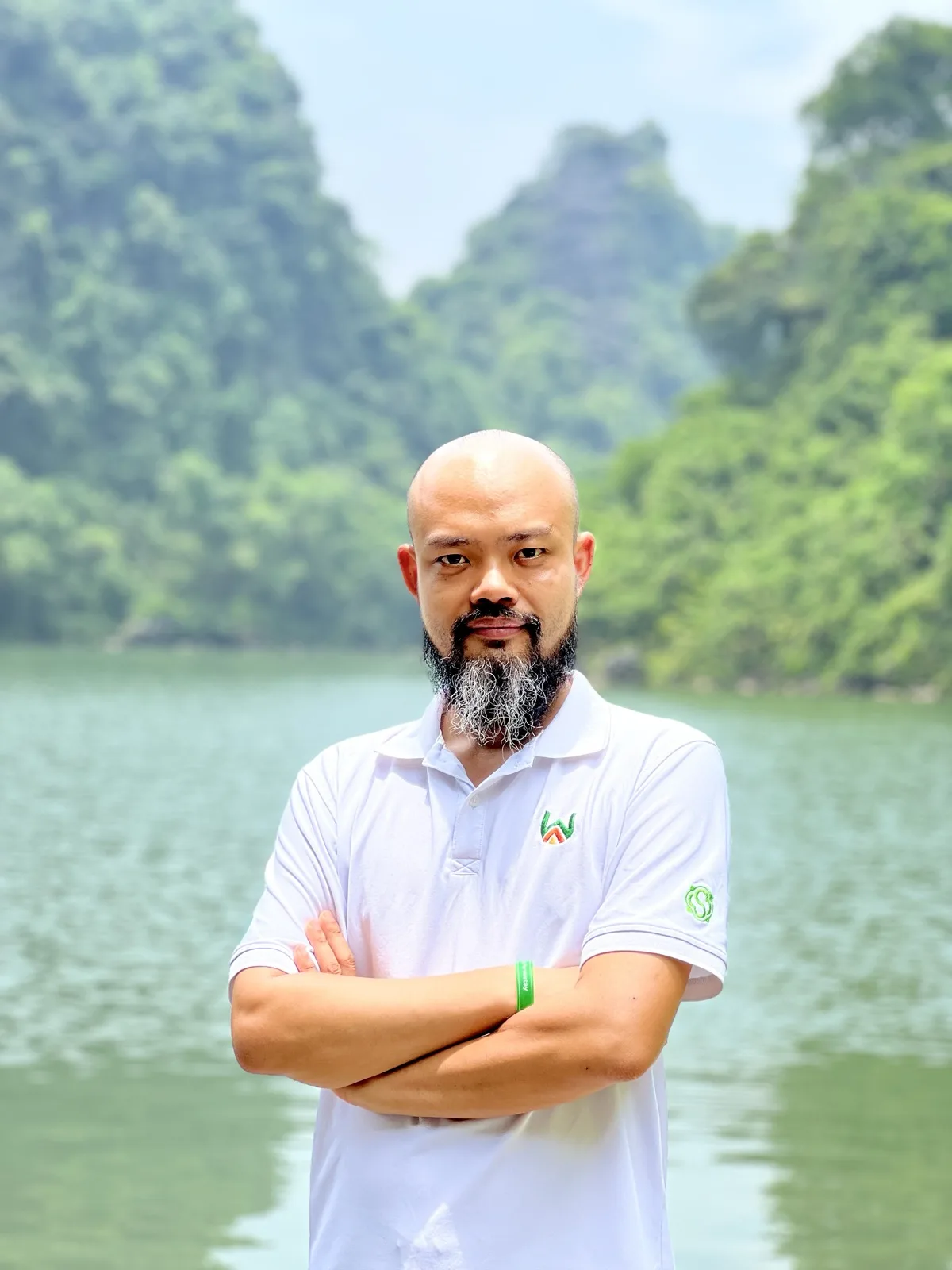 Nhà Hoạch Định Phạm Thanh Tùng đứng khoanh tay bên hồ nước, phía sau là dãy núi đá vôi phủ cây xanh