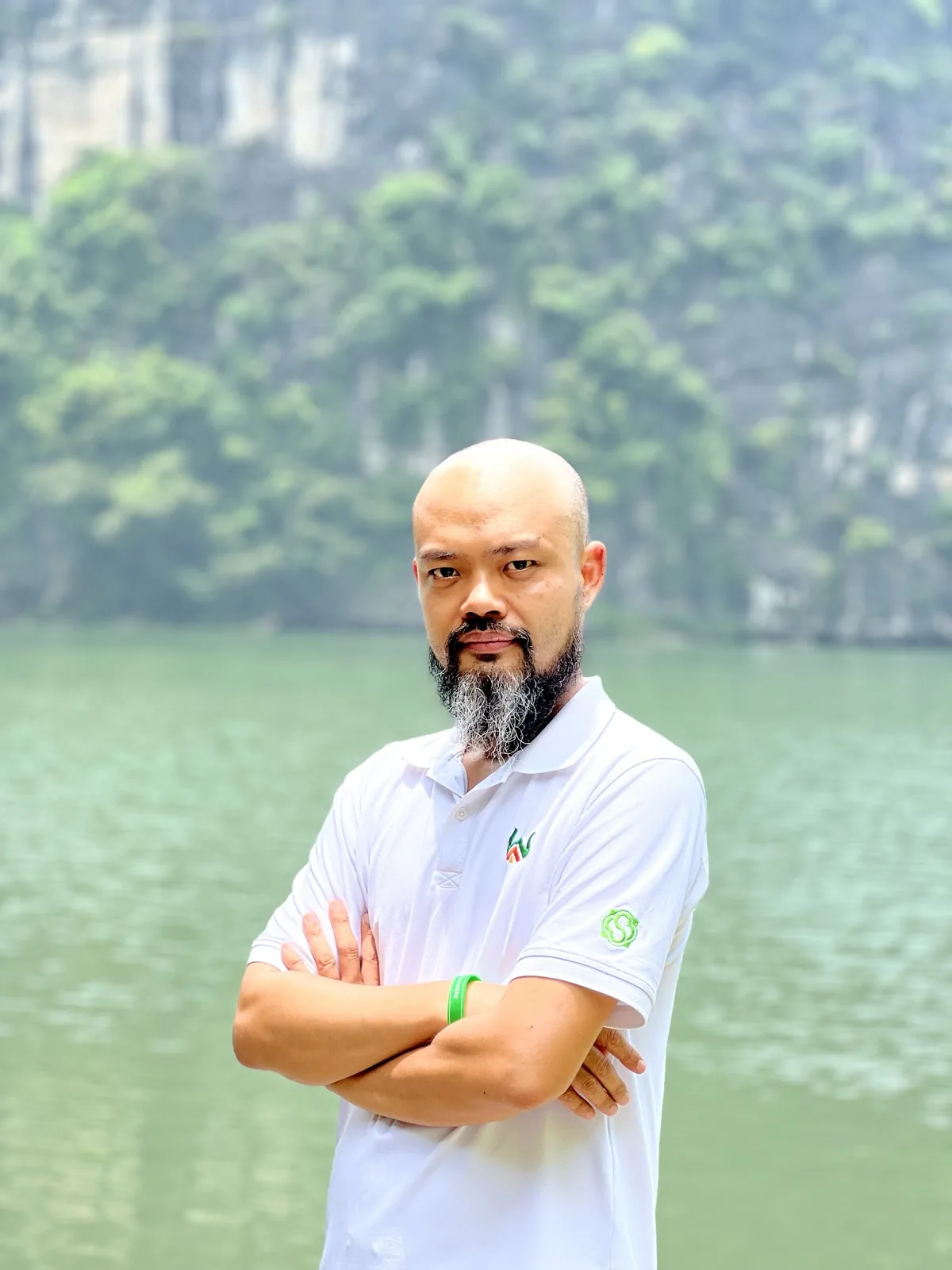 Nhà Hoạch Định Phạm Thanh Tùng đứng khoanh tay bên hồ nước, phía sau là dãy núi đá vôi phủ cây xanh