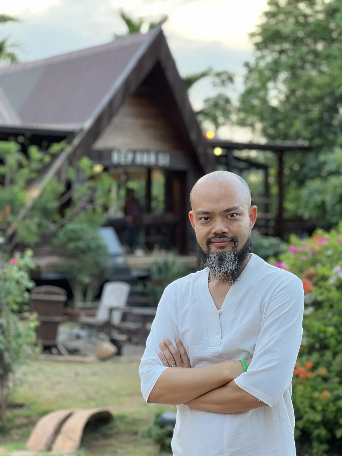 Phạm Thanh Tùng khoanh tay trước nếp nhà gỗ giữa vườn cây của một farmstay